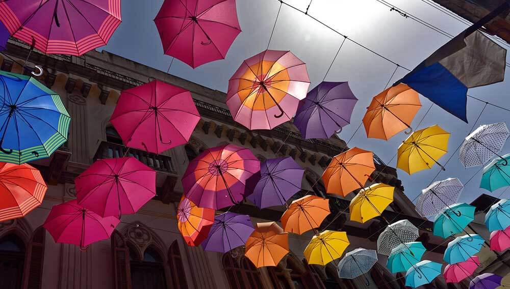 Viele bunte Schirme sind über einer Strasse zwischen Häusern aufgehängt. Die Sonne scheint und zwischen den Schirmen ist der Himmel zu sehen.