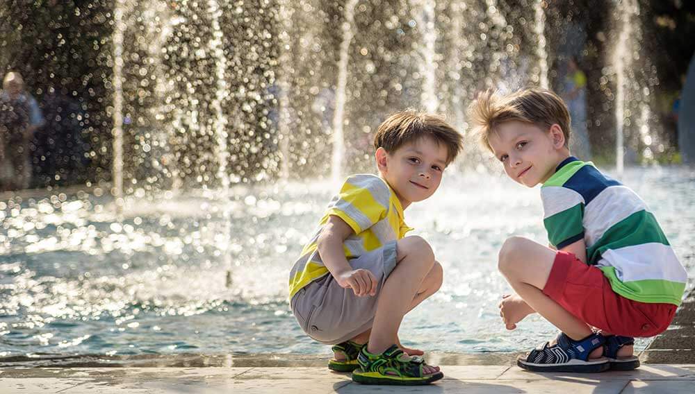 Zwei Buben spielen vor einem großen Springbrunnen. Illustration für den Geschäftsbereich Sommerkomfort der Firma Weatherpark.